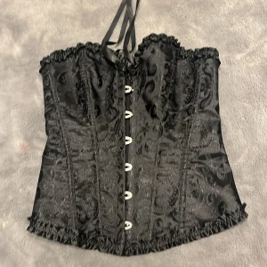 Svart korsett med snörning - Snygg svart korsett med dekorativt mönster och knappar framtill. Den har snörning bak för justerbar passform och volangdetaljer längs kanterna. Perfekt för en elegant look.