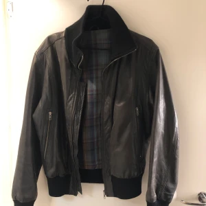 Svart skinnjacka från Bruuns Bazaar - Snygg svart skinnjacka från Bruuns Bazaar med dragkedja och ribbade muddar. Jackan har en klassisk design med en rutig insida i blå och bruna toner. Perfekt för en stilren look.