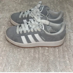 Grå Adidas Campus sneakers - Snygga grå Adidas Campus sneakers med klassiska vita ränder och snörning. Perfekta för en avslappnad stil. Skorna har en bekväm passform och är tillverkade i mocka för en stilren look.