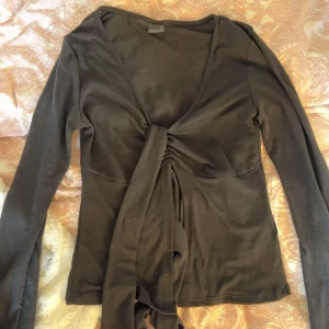 Svart långärmad topp med knyt - Snygg svart topp med långa ärmar och knyt framtill. Perfekt för en stilren look. Toppen har en v-ringad design. Aldrig använd, stl S från Gina!