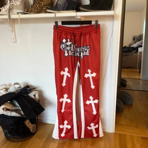 Röda byxor med korsmönster - Unika röda byxor med vita kors och textdetalj. Byxorna har en loose passform och elastisk midja för extra komfort. Perfekta för dig som vill sticka ut med en djärv stil.