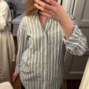 Randig skjorta från Ralph Lauren - Säljer en klassisk skjorta från Ralph Lauren i blått och gult med vertikala ränder. Skjortan har långa ärmar och knappar framtill. Perfekt för en stilren look.