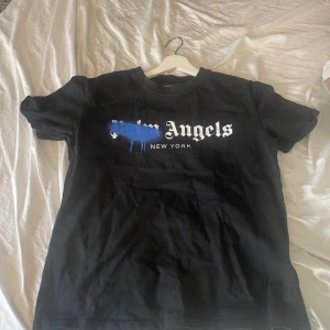 Svart t-shirt från Palm Angels - Säljer en svart t-shirt från Palm Angels med tryck i vitt och blått på framsidan. T-shirten har korta ärmar och en klassisk rund halsringning. Perfekt för en avslappnad stil.