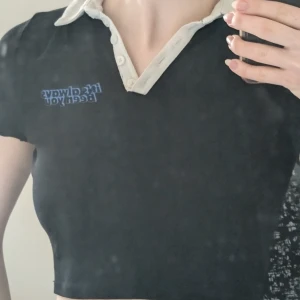 Svart croppad polotopp - Snygg svart croppad polotopp med vit krage och knappar. Perfekt för en trendig look. Textdetalj i blått på bröstet ger en cool touch.