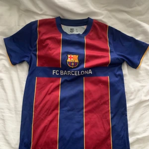 FC Barcelona fotbollströja - Säljer en klassisk FC Barcelona fotbollströja i blått och rött med gula detaljer. Tröjan har korta ärmar och klubbens emblem på bröstet. Perfekt för alla Barça-fans! Passar också XS