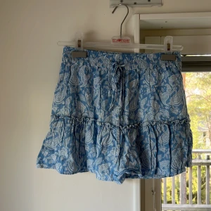 Blå mönstrad kjol från Zara - Säljer en blå kjol från Zara med ett vitt paisleymönster. Den har även inner shorts! Kjolen har en volangdetalj och knytband i midjan för en justerbar passform. Perfekt för en somrig look! Kjolen är väl omhändertagen men har några få lösa trådar 