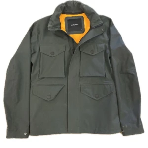 Grön jacka från Peak Performance - Säljer en stilren grön jacka från Peak Performance med orange foder. Jackan har flera praktiska fickor med knappar och en dragkedja framtill. Perfekt för utomhusaktiviteter.