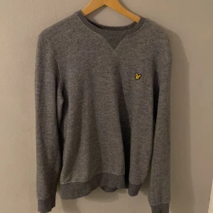 Grå tröja från Lyle & Scott - Säljer en stilren grå tröja från Lyle & Scott med deras ikoniska logga på bröstet. Tröjan har långa ärmar och en rund halsringning. Perfekt för en avslappnad look.
