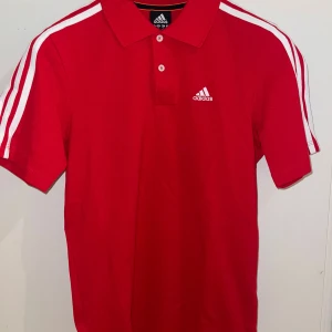 Röd pikétröja från Adidas - Säljer en snygg röd pikétröja från Adidas med klassiska vita ränder på axlarna och en broderad logga på bröstet. Perfekt för en sportig look! 👟Diverse andra nya piketröjor. 179kr st olika st. 