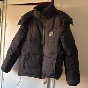 Svart pufferjacka från Trapstar - Säljer en svart pufferjacka från Trapstar med broderad logga på ryggen. Jackan har en dragkedja framtill och en stor huva. Perfekt för kalla dagar med sin vadderade design.