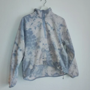 Blå och vit fleece från Hollister - Mysig blå och vit fleece från Hollister med ett snyggt batikmönster. Tröjan har en halv dragkedja framtill och en hög krage för extra värme. Perfekt för kyliga dagar!