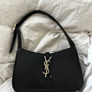 Svart axelväska från Saint Laurent - Snygg svart axelväska från Saint Laurent i skinn med det ikoniska YSL-emblemet i silver på framsidan. Väskan har en justerbar axelrem med ett elegant spänne i guld. Perfekt för att ge en lyxig touch till din outfit.