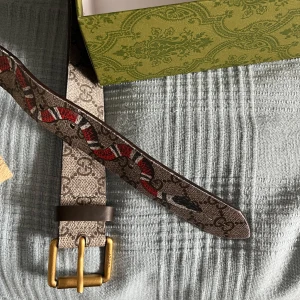 Mönstrat bälte från Gucci - Säljer ett stilrent bälte från Gucci med ett unikt mönster och en iögonfallande orm i rött och vitt. Bältet har en klassisk spänne i guld och är perfekt för att ge din outfit en lyxig touch.