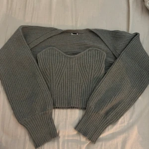 Grå ribbad stickad topp - Säljer en stilren grå ribbad stickad topp med matchande bolerotopp. Toppen har en hjärtformad urringning och bolerotoppen har långa ärmar. Lite ”trasig” vid kragen se sista bild. Köpt så, oanvänd. 