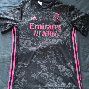 Real Madrid tredjeställ 20-21 - Säljer min Real Madrid tröja från säsongen 20-21. Snygg svart fotbollströja med grått mönster och rosa detaljer från Adidas. Tröjan har korta ärmar och klubbmärke på bröstet. Perfekt för fotbollsfans som vill sticka ut på planen.