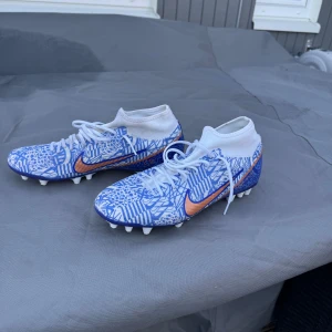 Nike fotbollsskor med blått mönster - Snygga fotbollsskor från Nike med ett unikt blått och vitt mönster. Skorna har en hög ankelkrage för extra stöd och en orange Nike-logga på sidan. Perfekta för fotbollsplanen! Knappt använda och ät i mycket bra form
