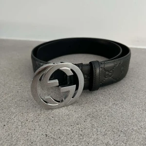 Gucci bälte 85cm - Snyggt svart skärp från Gucci med ett stort, runt silverfärgat spänne. Skärpet har ett diskret mönster med märkets logotyp präglat längs hela remmen. Perfekt för att ge en lyxig touch till din outfit.