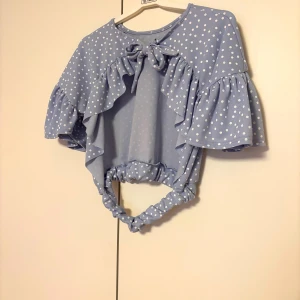 Blå prickig topp med volanger - Blå topp med vita prickar och volangdetaljer. korta ärmar och en söt knytning bak med öppen rygg . Perfekt till sommaren!