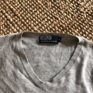 Grå tröja från Polo Ralph Lauren - Säljer en stilren grå tröja från Polo Ralph Lauren med v-ringning och broderad logotyp på bröstet. Tröjan har långa ärmar och är perfekt för en avslappnad men ändå elegant look. Den är storlek M men den är rätt liten i storleken så skulle säga att den passar bättre på S. Hör av er vid fler frågor eller funderingar!😁