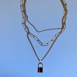 Guldhalsband med hänglås - Snyggt guldhalsband med flera kedjor och ett hänglås som detalj. Kedjorna har olika länkar vilket ger en trendig lager-på-lager-look. Perfekt för att ge din outfit en edgy touch.
