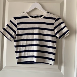 Randig kortärmad topp från Zara - Säljer en snygg randig magtröja/topp från Zara i vitt och blått. Den har korta ärmar och en croppad passform, perfekt för en avslappnad stil. Toppen är tillverkad i ett mjukt material som känns skönt mot huden.