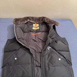 Svart dunväst från Para Jumpers - Säljer en svart dunväst från Para Jumpers med dragkedja och knappar. Västen har en mjuk krage i pälsimitation och praktiska fickor med dragkedja. Perfekt för kyligare dagar.