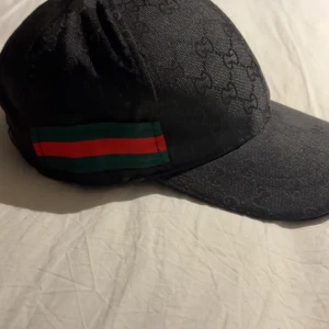 Svart keps med mönster och randig detalj - Snygg svart keps med diskret mönster och en randig detalj i grönt och rött på sidan. Perfekt för en stilren look.