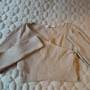 Beige ribbad omlottopp från NA-KD i storlekS - Säljer en snygg beige ribbad omlottopp från NA-KD. Toppen har en v-ringad design och långa ärmar, perfekt för en stilren look. Den är tillverkad i ett mjukt och bekvämt material.