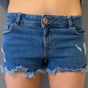 Blå jeansshorts med fransar - Säljer ett par snygga blå jeansshorts med fransiga kanter. De har en klassisk femficksdesign och en knapp framtill. Perfekta för sommardagar!