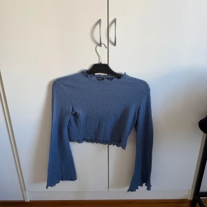 Blå ribbad tröja - Säljer en snygg blå ribbad tröja med långa ärmar och en croppad design. Tröjan har en vågig kant vid ärmslut och nederkant som ger en extra touch. Perfekt för en stilren look!