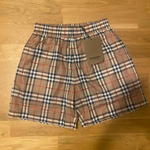 Rutig shorts från Burberry - Snygga rutiga shorts från Burberry i klassiskt mönster med beige, blå och röda rutor. De har en elastisk midja för bekväm passform. Perfekta för en stilren look.