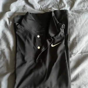 Svart pikétröja från Nike - Säljer en svart pikétröja från Nike med Dri-FIT-teknologi. Tröjan har en klassisk krage med knappar och det ikoniska Nike-logotypen på bröstet. Perfekt för en sportig look eller en avslappnad dag. Stretch material. Golf pike eller bara en vanlig snygg piké 
