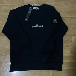 Svart tröja från Stone Island - Säljer en svart tröja från Stone Island med deras ikoniska logga på bröstet och en stor logga på ryggen. Tröjan har långa ärmar och en rund halsringning. Perfekt för en stilren look.