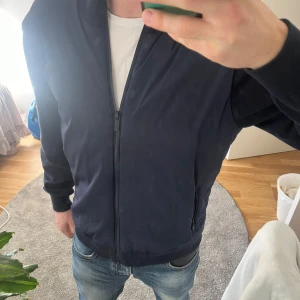 Mörkblå John Henric cardigan  - Säljer en stilren mörkblå cardigan med dragkedja framtill. Jackan har långa ärmar och ribbade muddar vid ärmslut och nederkant. Perfekt till våren för en casual look.