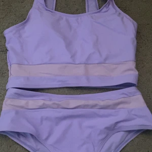 Lila bikini med breda axelband - Snygg lila bikini med breda axelband och kontrasterande ljusare banddetalj. Perfekt för en dag på stranden eller vid poolen. Toppen har en sportig design och trosorna har en bekväm passform.