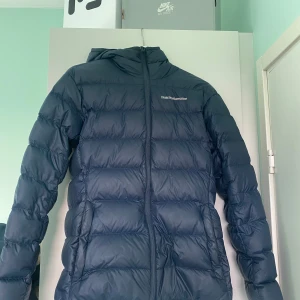 Mörkblå dunjacka från Peak Performance - Säljer en snygg mörkblå dunjacka från Peak Performance. Jackan har en dragkedja framtill och är långärmad med en quiltad design. Perfekt för kyligare väder med sin varma och bekväma passform.