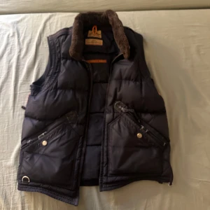 Svart dunväst från Parajumpers - Säljer en svart dunväst från Parajumpers Masterpiece Series. Västen har en mysig pälsfodrad krage och flera praktiska fickor med dragkedja och knappar. Perfekt för kyliga dagar med sin quiltade design och robusta känsla. Passar S oxå men mest som M