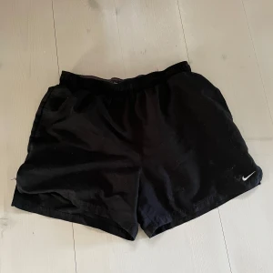 Svarta träningsshorts från Nike - Säljer ett par svarta träningsshorts från Nike. De har en elastisk midja och är perfekta för sport och träning. Nike-loggan syns diskret på ena benet. Perfekta för en aktiv livsstil!