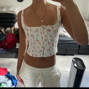 Blommig korsettopp med knytning - Säljer en söt blommig korsettopp med knytning framtill. Toppen är vit med rosa och gröna blommor och har en croppad passform. Perfwkr nu till sommar, köp via köp nu❤️