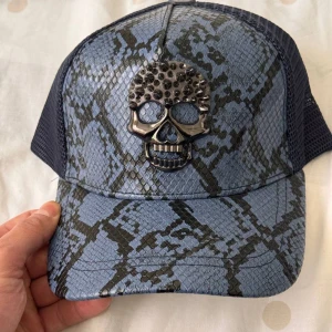 Pp keps - Snygg blå keps med ett coolt dödskallemönster i relief på framsidan. Kepsen har ett nätmaterial på baksidan för bättre ventilation och en justerbar rem för perfekt passform. Perfekt för att ge din outfit en edgy touch!