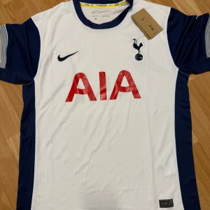Vit och marinblå fotbollströja från Nike - Säljer en vit och marinblå fotbollströja från Nike med korta ärmar. Tröjan har en röd AIA-logga på framsidan och ett Tottenham Hotspur-märke. Tillverkad i Dri-FIT-material för optimal komfort. Perfekt för fotbollsfans!