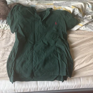 Mörkgrön pikétröja från Ralph Lauren - Snygg mörkgrön pikétröja från Polo Ralph Lauren med klassisk krage och korta ärmar. Tröjan har den ikoniska röda broderade loggan på bröstet och är gjord i mjuk bomull. Perfekt för en stilren och avslappnad look.{ använd ett par gånger}