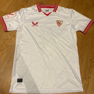 Sevilla FC Sergio Ramos tröja Castore - Vit Sevilla FC matchtröja från Castore med röda detaljer och klubbmärke på bröstet. Kortärmad med röd/vit randig krage och ärmslut. På ryggen står det 'Sergio Ramos' och nummer 4 i rött. Tillverkad i lätt och ventilerande material.