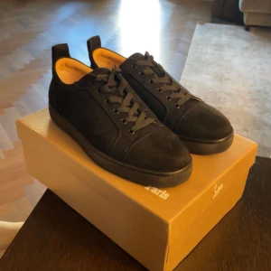 Svarta Louboutin, strl 43 - Snygga svarta sneakers från Louboutin med klassisk röd sula och svarta snören. Ovandel i mocka och skinn, insida i beige läder. Perfekt för dig som vill sticka ut med exklusiva detaljer och stilren design.