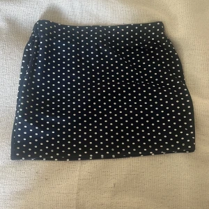 Svart prickig kortkjol - Snygg mörkblå kortkjol med vita prickar, perfekt för dig som gillar en klassisk men ändå lekfull stil. Kjolen har en rak tajt passform och är superenkel att matcha med både t-shirt och stickad tröja. Prickmönstret ger en retrovibe som aldrig går ur tiden.