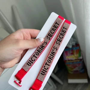 Victoria's Secret glittriga bh band  - Två snygga bh band från Victoria's Secret i starkt rosa med glittrande stenar och texten 'VICTORIA'S SECRET' i svart. Armbanden har justerbar längd och är perfekta för att ge din outfit extra bling. Materialet är textilband med glittriga detaljer.