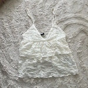 Vit volang-linnetopp från Gina Tricot - Supersöt vit linnetopp från Gina Tricot med tunna axelband och volanglager. Toppen har v-ringning och är tillverkad i ett lätt, krinklat tyg som ger en luftig känsla. Perfekt för varma dagar och enkel att styla med jeans eller kjol.