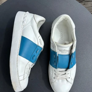 Valentino Garavani sneakers vit/blå 38 - Snygga Valentino Garavani sneakers i vitt skinn med bred blå läderdetalj över mitten. Klassisk rund tå, vita snören och platt sula med ikoniska nitar på hälen. Perfekta för dig som vill sticka ut med en lyxig touch.