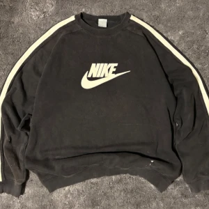 Vintage Svart Nike sweatshirt med vita ränder - Svart sweatshirt från Nike med klassisk logga framtill och vita ränder längs ärmarna. Tröjan har en loose passform, perfekt för en sportig och avslappnad stil. Det står stl XL på etiketten men den hade kunnat vara L. Det finns lite färgfläckar (se bild)