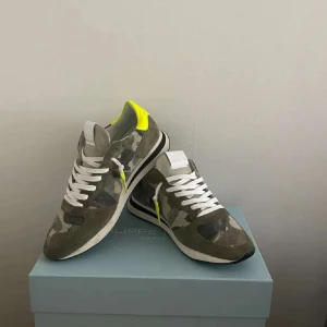Philippe Model sneakers camo och neon - Snygga Philippe Model sneakers i camouflage-mönster med detaljer i neon-gult och vita partier. Skorna har vita snören, rund tå och platt sula. Materialmix av mocka och syntet, perfekt för dig som gillar streetstyle med en twist. Kan gå ner lite i pris 
